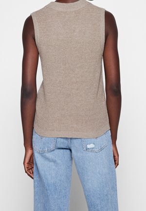 Personne portant un gilet tricoté beige sans manches et un jean taille haute bleu clair avec des poches arrière, vue de dos sur un fond uni.