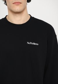 Crni dukserica s okruglim izrezom, s bijelim izvezenim tekstom "NoProblemo" na prsima. Glatka, jednobojna tekstura, ležeran dizajn.