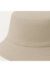 Beige emmerhoed gemaakt van textuurstof, met een brede, platte rand en gestikte accenten langs de randen.