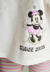 Weißes T-Shirt mit einem Grafikdesign eines Cartoon-Maus in einem rosa gepunkteten Rock und gelben Schuhen, mit dem Text "SEIT 1928" darunter.