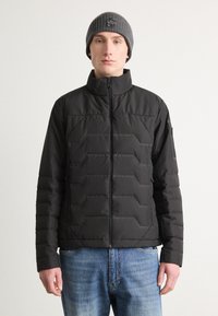 Crna quilted jakna s visokim ovratnikom i prednjim zatvaračem, sa teksturiranim uzorkom. Uparena s sivim pletenim šeširom minimalnog dizajna.