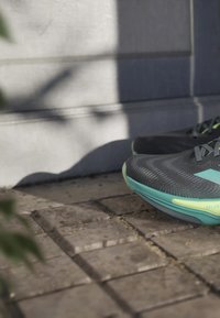 Pair of black and teal running shoes on stone pavement beside a shaded gray concrete wall.