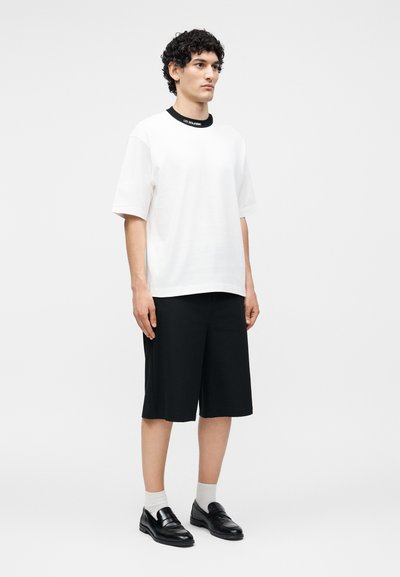 Νέος άνδρας με σγουρά μαλλιά που φοράει λευκό φαρδύ μπλουζάκι, μαύρα φαρδιά σορτς, λευκές κάλτσες και μαύρα δερμάτινα μοκασίνια, στέκεται μπροστά σε απλό φόντο.