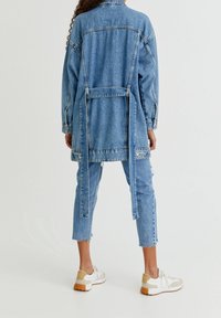 Veste en jean à coupe ample, de couleur bleu clair, manches longues, ceinture nouée à la taille, et pantalon en jean déchiré assorti à des baskets blanches.