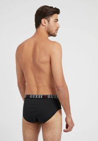 Schwarzer Baumwoll-Slip mit breitem Bund, der ein kontrastierendes "GUESS"-Logo zeigt. Glatte Textur, figurbetontes Design und klassische Silhouette.