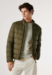 Veste olive verte rembourrée avec un col haut, fermeture éclair et texture matelassée. Portée sur un pull gris clair et un pantalon crème.