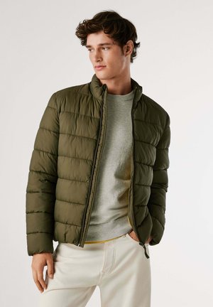 Chaqueta acolchada de color verde olivo con un cuello alto, cierre de cremallera y textura acolchada. Lleva puesta sobre un suéter gris claro y pantalones crema.