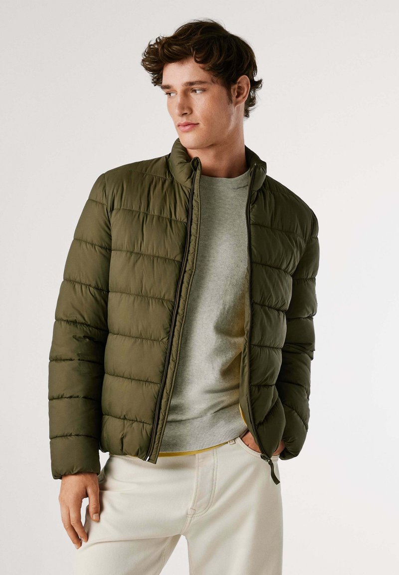 Veste olive verte rembourrée avec un col haut, fermeture éclair et texture matelassée. Portée sur un pull gris clair et un pantalon crème.