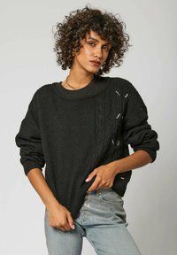 Pull en maille noir avec un motif en torsades, coupe oversize et manches retroussées, porté sur un haut foncé, associé à un jean en denim clair.
