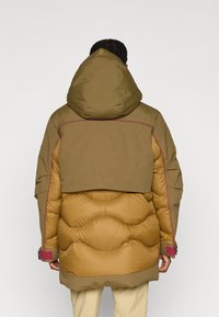 Kurtka puchowa w kolorze brązowym i musztardowym, z kapturem, dużymi pikowanymi sekcjami oraz kontrastującym różowym przeszyciem wzdłuż szwów.