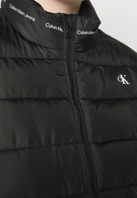 Černá prošívaná bunda s vysokým límcem, předním zipem a logem "Calvin Klein Jeans" na límci. Hladký, lesklý povrch.