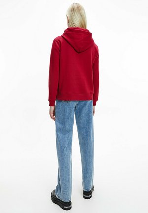 Sweat à capuche rouge avec une grande capuche, des poignets et un ourlet côtelés, associé à un jean bleu clair à jambes larges et des chaussures noires à semelles épaisses.