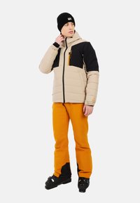 Gevoerde beige en zwarte jas met oranje accenten, gecombineerd met oranje broek. Model draagt een zwarte beanie en zwarte schoenen.