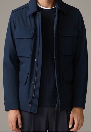 Mężczyzna w granatowej kurtce z dużymi klapowymi kieszeniami, nałożonej na ciemny, teksturowany sweter i ciemne spodnie, na jednolitym tle.