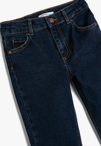 Jeans de mezclilla azul oscuro de corte ajustado con costuras naranjas, cierre de botón y bolsillos. Textura suave, diseño clásico de cinco bolsillos.