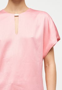 Gros plan sur une personne portant un haut à manches courtes texturé rose pâle avec une encolure fendue étroite maintenue par une petite boucle métallique rectangulaire.