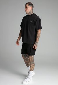 Camiseta de algodón negra de manga corta, combinada con pantalones cortos cargo negros. Las características de estilo incluyen tatuajes y zapatillas blancas.