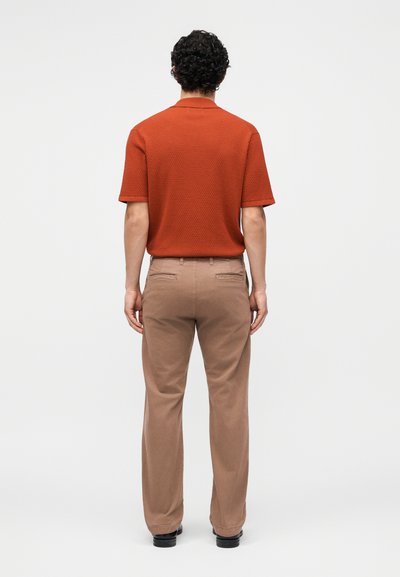 Homme aux cheveux bouclés, debout de dos, portant une chemise texturée manches courtes couleur rouille et un pantalon beige avec des chaussures noires.