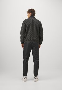 Giacca nera a fantasia con stampa geometrica, polsini e orlo elastici. Abbinata a pantaloni neri e sneakers bianche. Vista posteriore.