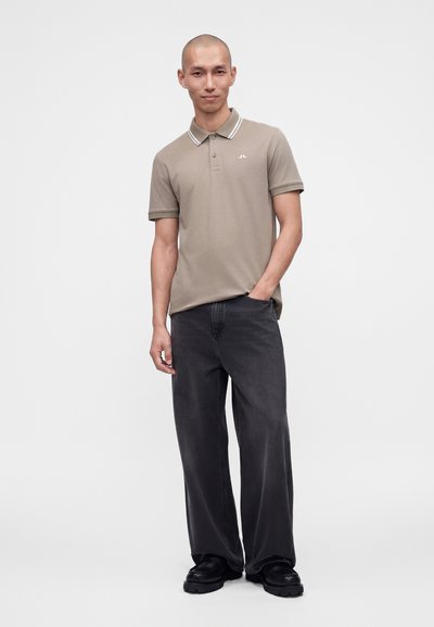 Homme portant un polo taupe et un jean noir ample avec des chaussures noires, debout avec une main dans la poche devant un fond uni.