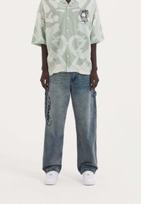 Light green short-sleeve shirt with graphic print, buttoned front. Blue baggy jeans with black lettering on the leg. White sneakers visible.