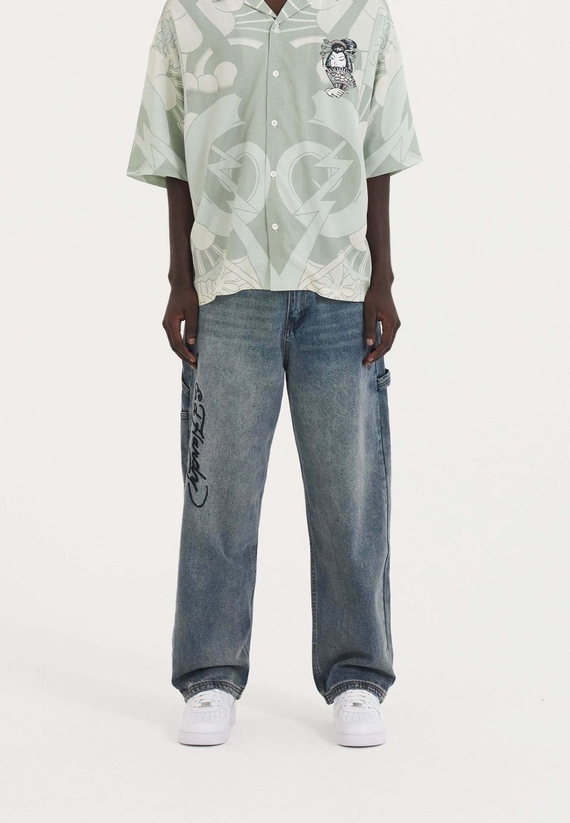 Ljust grönt kortärmat skjorta med grafiskt tryck, knäppt fram. Blå baggy jeans med svart text på benet. Vita sneakers synliga.