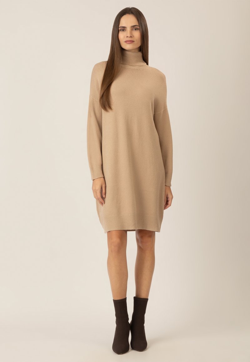 Beige Rollkragenpullover-Kleid mit langen Ärmeln, gerippten Bündchen und Saum, das eine lockere Form und eine glatte Textur hat. Kombiniert mit dunkelbraunen Knöchelsocken.