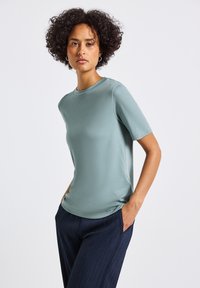 Žena s kudrnatými vlasy, oblečená v světle zeleném tričku s krátkým rukávem a tmavých plisovaných kalhotách, stojící s rukama v kapsách na bílém pozadí.