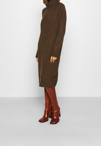 Robe col roulé en tissu tricoté marron foncé, longueur au genou, avec des poignets côtelés. Associée à des bottes à talons hautes brillantes rouge-brun.