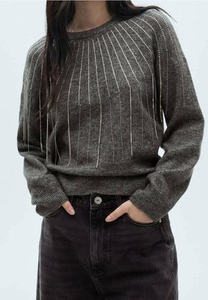 Kvinde iført brun strikket sweater med lodrette sølvperlelinjer kombineret med sorte højhælede jeans, hænderne stukket i forlommerne.