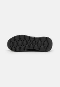 Suela de zapatilla negra con una superficie de goma texturizada, con agarres en forma de óvalo y patrones elevados para mayor tracción. Bordes suaves con un diseño curvado.