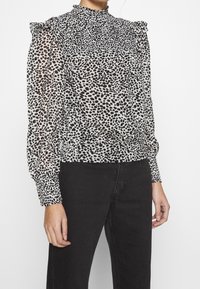 Transparent blus med hög krage och volangiga axlar; har ett svartvitt prickigt mönster. Elastiska manschetter och nederkant.