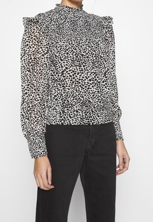 Blusa trasparente con collo alto e spalle arricciate; presenta un motivo a pois bianco e nero. Polsini ed orlo inferiore elastici.