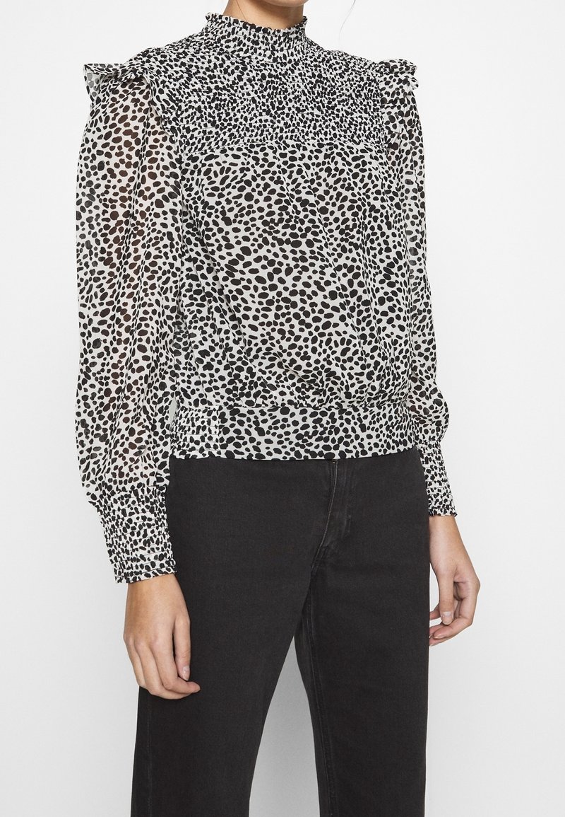 Transparent blus med hög krage och volangiga axlar; har ett svartvitt prickigt mönster. Elastiska manschetter och nederkant.