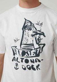 Camiseta blanca con un diseño gráfico negro de una figura con sombrero, acompañada de aves y el texto "Altona-Dock" junto con un ancla.