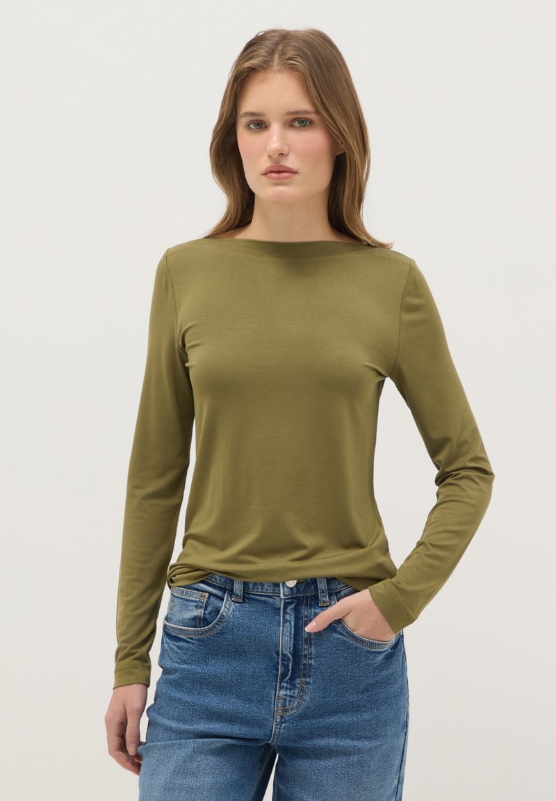 Frau steht mit einer Hand in der Tasche, trägt ein olivgrünes, langärmliges Oberteil und blaue Jeans vor einem einfarbigen Hintergrund.