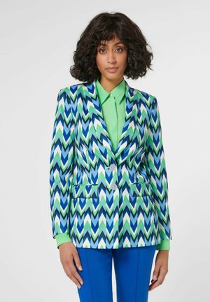 Frau mit schulterlangem, lockigem Haar trägt ein lebhaftes, zickzackmusteres Blazer in Blau, Grün, Schwarz und Weiß über einem hellgrünen Hemd und blauen Hosen.