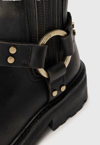 Botín negro de cuero a la altura del tobillo con costuras laterales, acentos metálicos y una correa decorativa con una hebilla circular. Suela gruesa con patrón de tracción.