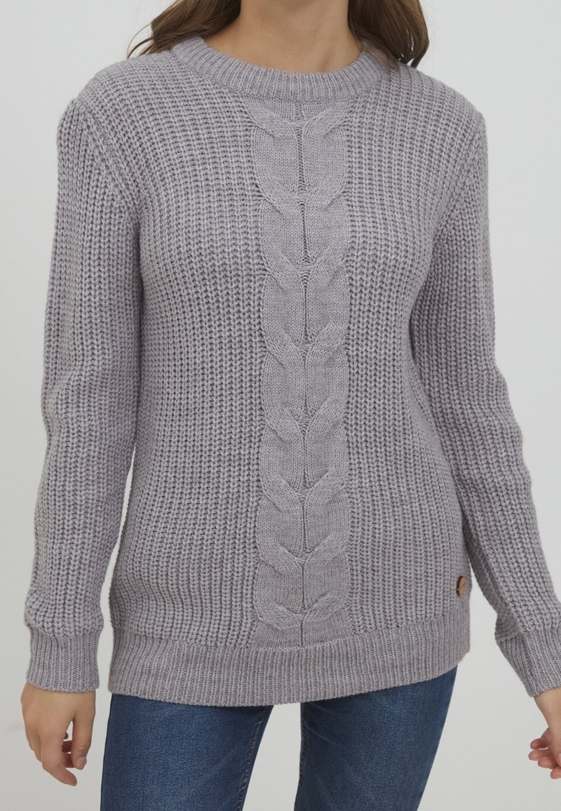 Femme portant un pull en tricot gris clair avec un motif torsadé central et un point texturé, associé à un jean bleu, sur un fond uni.