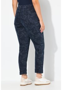 Tmavě modré džíny s jemným vzorem růží, elastickým pasem a zadními kapsami, kombinované s bílými teniskami na světlém pozadí.