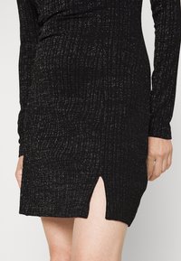 Schwarzes, geripptes Kleid aus strukturiertem Stoff, mit langen Ärmeln und einem seitlichen Schlitz bis zur Oberschenkelhöhe, verziert mit dezenten metallischen Flocken.