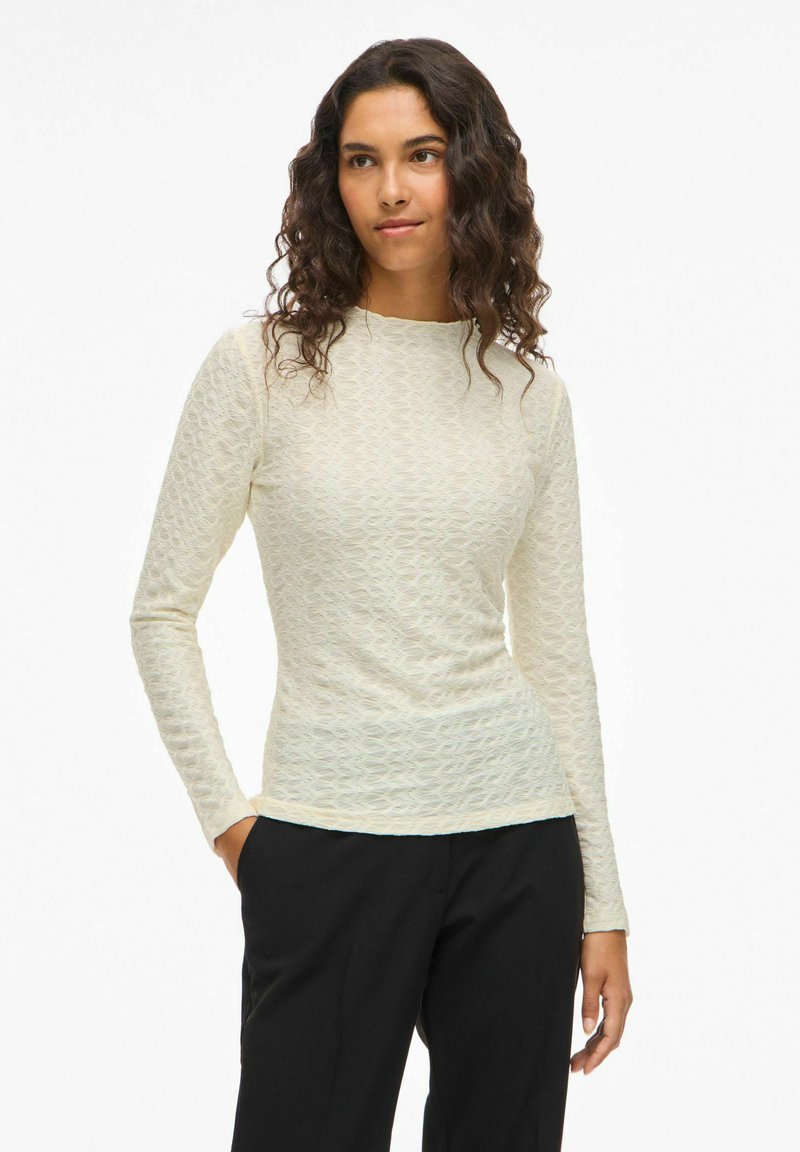 Langärmliges Oberteil in hellem Creme mit einem strukturierten Muster. Figurbetonter Schnitt mit Rundhalsausschnitt. Kombiniert mit schwarzen Hosen.