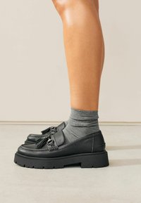 Mocasines de cuero negro con borlas y acentos metálicos, que presentan una suela gruesa y texturizada. Llevados con calcetines grises hasta el tobillo. Vista lateral.