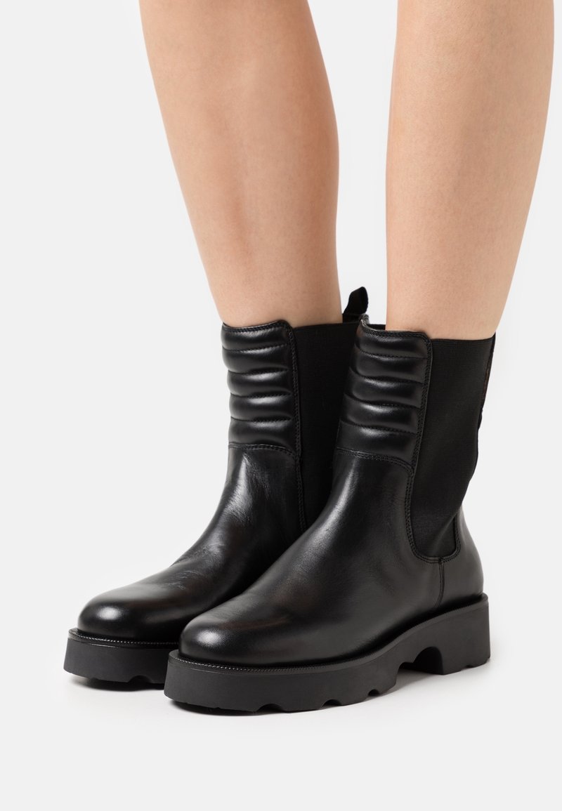 Bottes courtes en cuir noir avec des panneaux latéraux élastiques, des détails côtelés en haut, et une semelle noire épaisse avec un motif de bande de roulement.