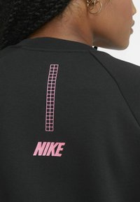 Svart sweatshirt med ett rosa rutmönster och "NIKE"-logotyp i rosa på baksidan. Tyget verkar mjukt med en texturerad finish.