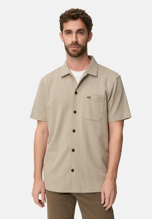 Homme aux cheveux courts bouclés et à la barbe, portant une chemise beige à manches courtes boutonnée et un pantalon marron, debout devant un fond uni.