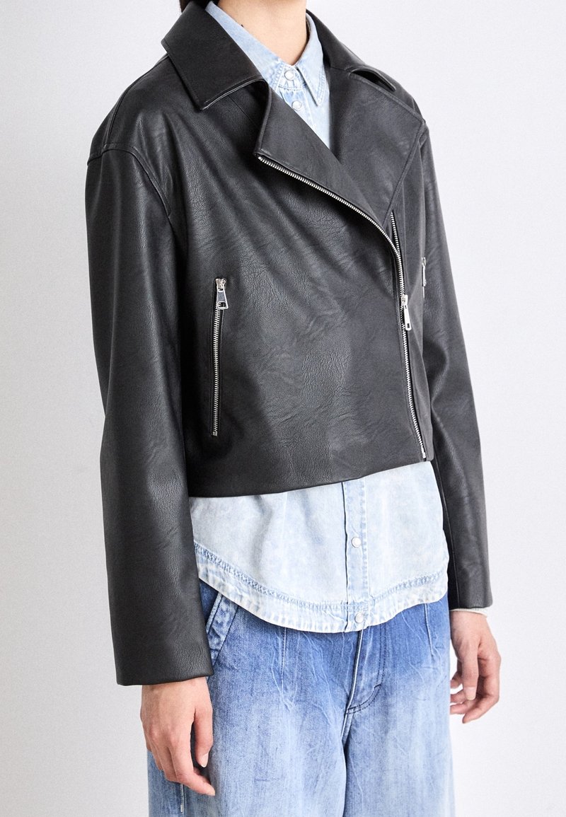 Giacca di pelle nera corta con colletto ampio, chiusura con zip e due tasche laterali con zip; abbinata a una camicia azzurra con bottoni.