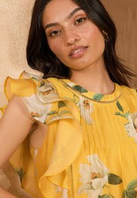 La blusa gialla a fiori presenta maniche arricciate e un design a pieghe. Il tessuto è morbido con motivi di foglie verdi e fiori bianchi.