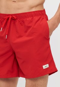 Punaiset uimapuvut, jotka on valmistettu kevyestä materiaalista. Niissä on elastinen vyötärö, jossa on nyöri sekä sivutaskut. Sisältää pienen logopatchin.