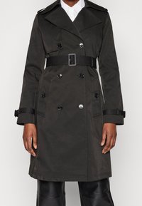 Mörkgrön trenchcoat med dubbelknäppt design, stor krage, bälte och knappdetaljer. Har sidofickor och manschettremmar.